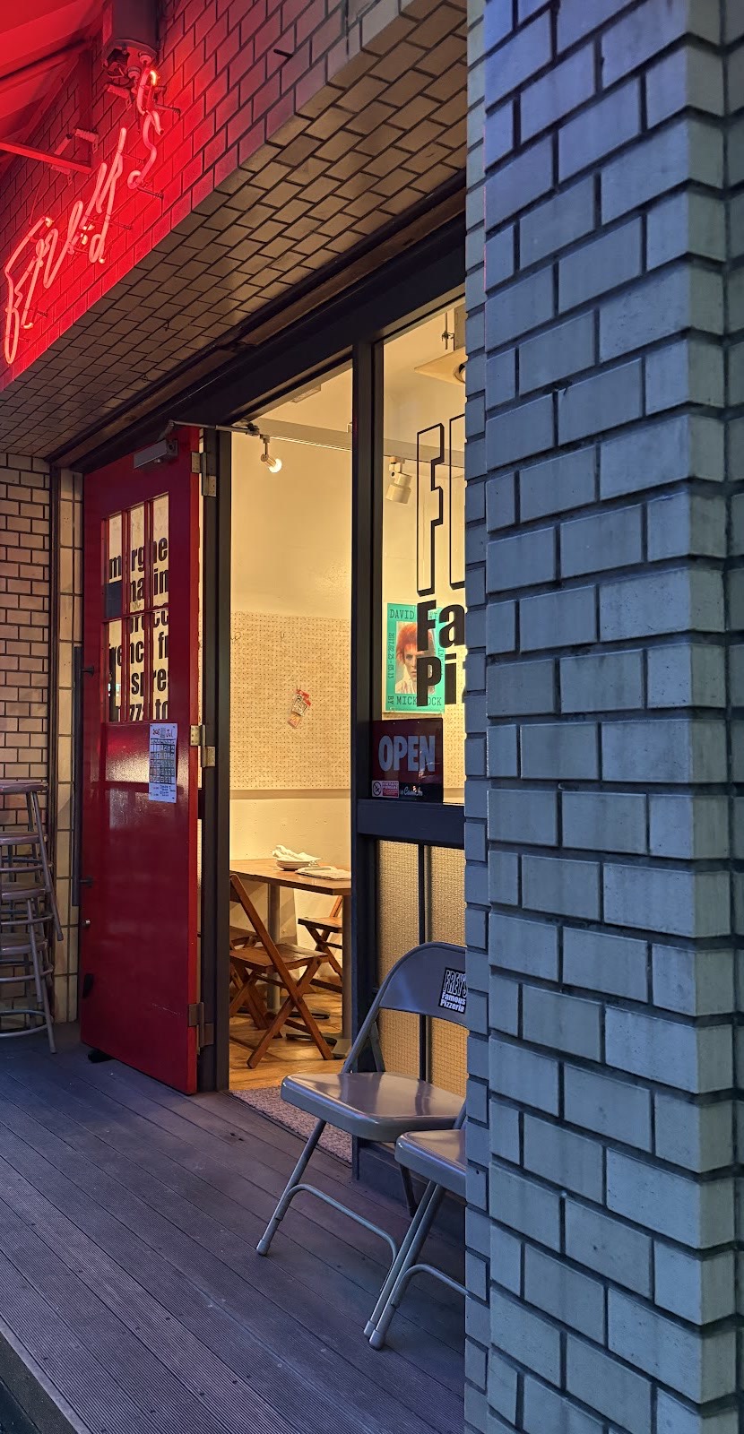 夕暮れの店先。赤いストライプの日よけの下に赤いネオンの「Frey's」、開いた赤い扉、白いタイルの壁、ウッドデッキに折りたたみ椅子。