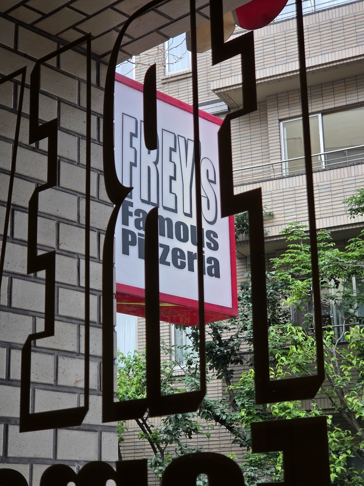窓の大きな縁取り文字越しに見える「FREY'S Famous Pizzeria」の突き出し看板。奥に木々とタイル張りの建物。