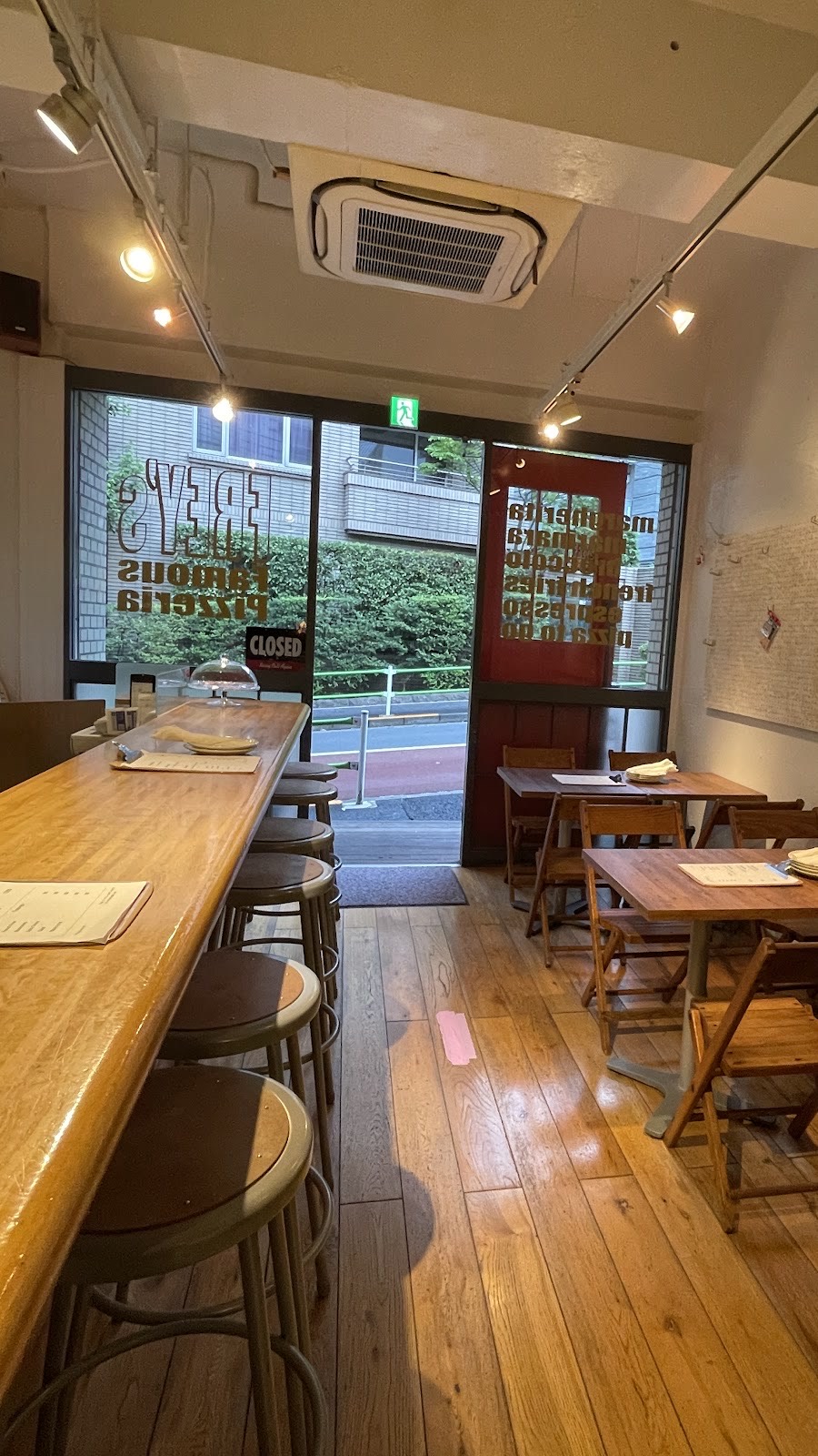 昼の店内から入口を望む。丸いスツールが並ぶ木のカウンター、木の折りたたみ椅子のテーブル席、窓の「FREY'S Famous Pizzeria」の文字（裏返し）と開いた赤い扉。