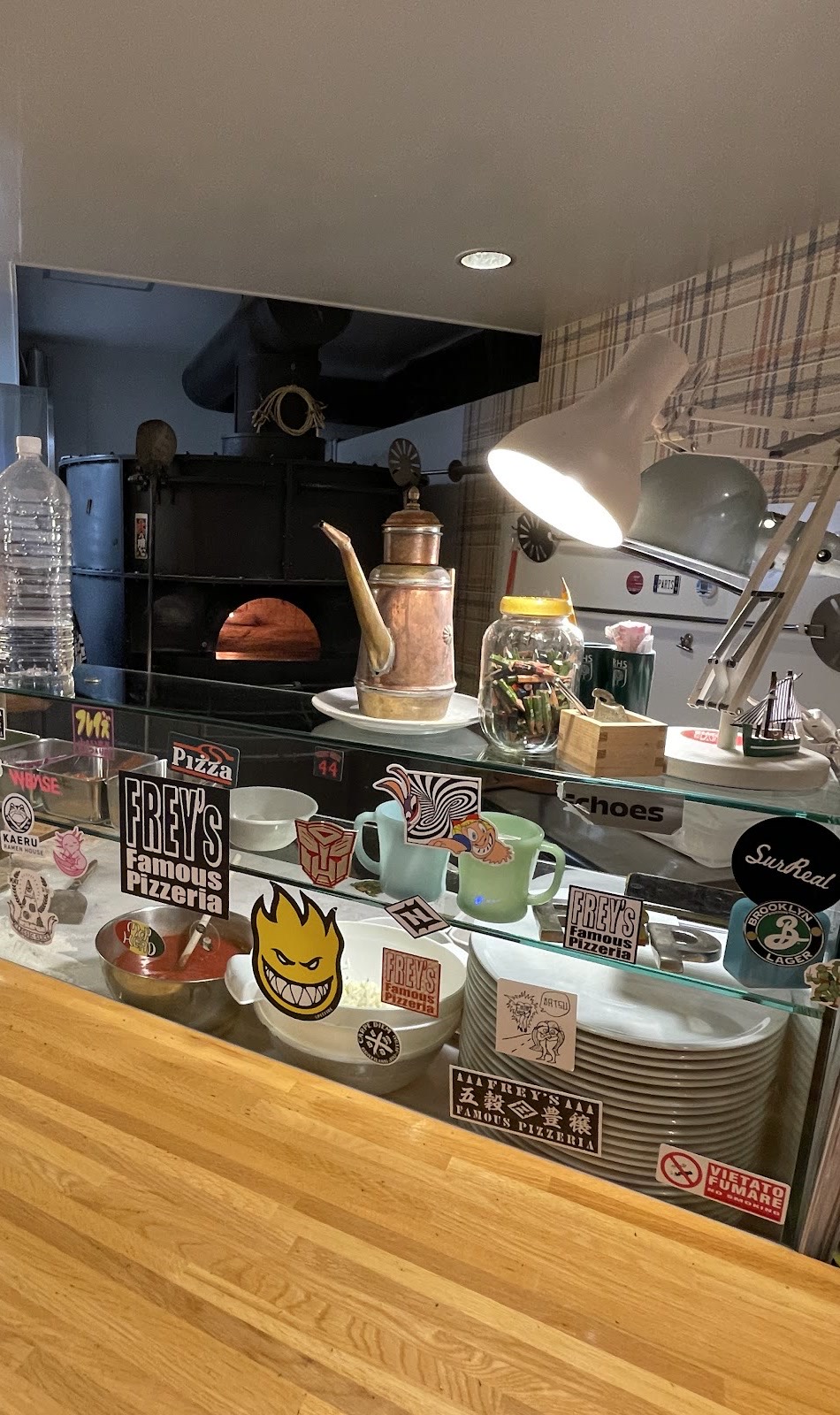 「FREY'S Famous Pizzeria」などのステッカーが貼られたガラスの食材ケース。その奥に火の灯る黒いドーム型の窯、銅のオイル差し、デスクライト。