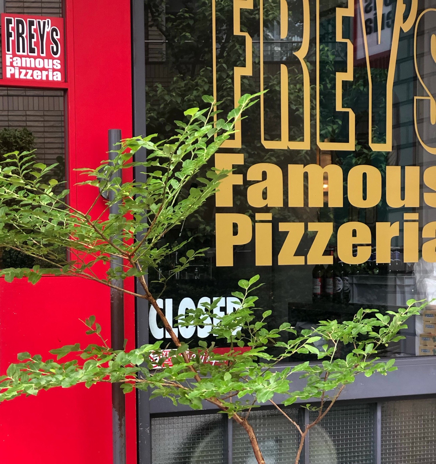 昼の店先。赤い扉に「FREY's Famous Pizzeria」のステッカー、窓に金色の「FREY'S Famous Pizzeria」の文字、手前に緑の枝。