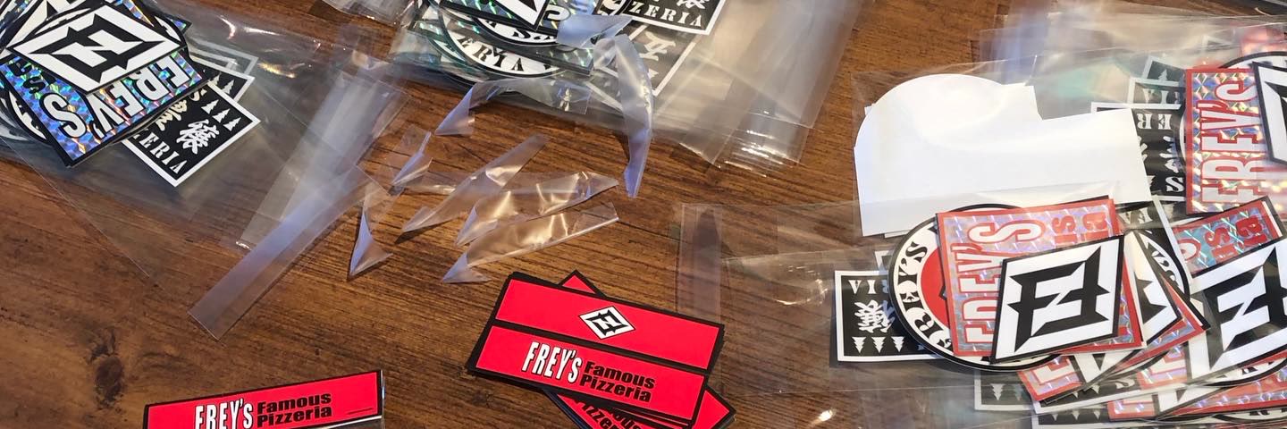 木のテーブルに並んだ、袋入りのFREY'Sのステッカー。菱形のFの紋や「FREY'S Famous Pizzeria」の赤い台紙。